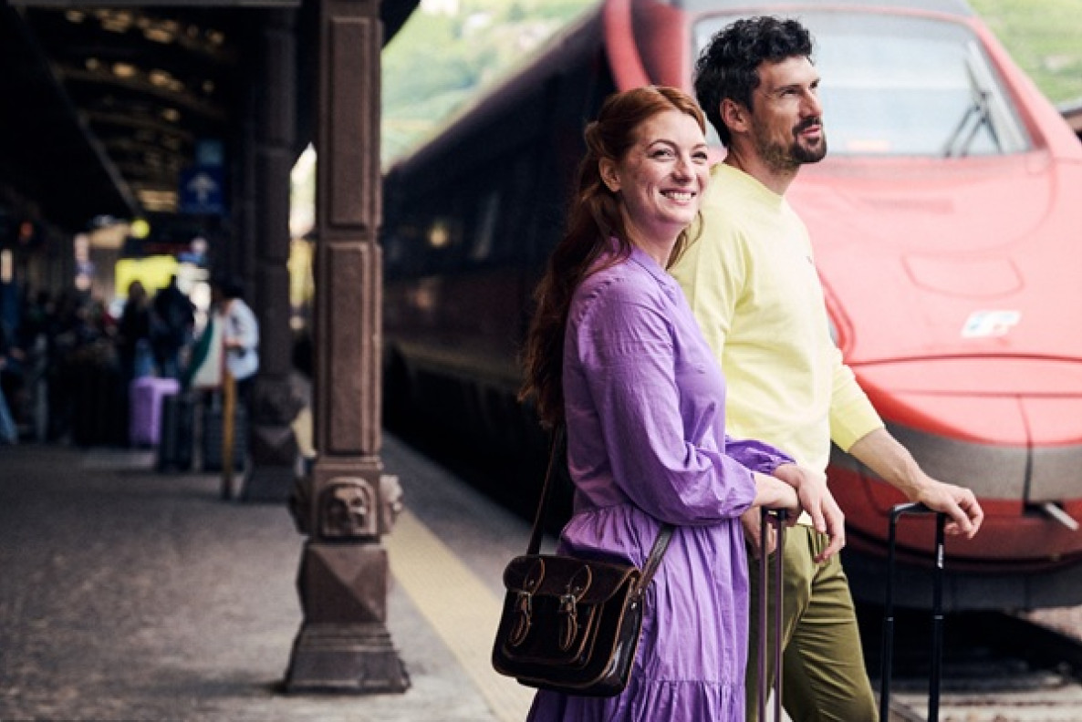 Arrivare a Bolzano in treno Coppia con valigie sul binario della stazione di Bolzano, davanti a un treno rosso