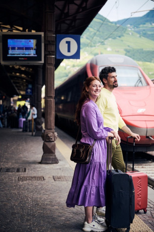 Arrivare a Bolzano in treno Coppia con valigie sul binario della stazione di Bolzano, davanti a un treno rosso