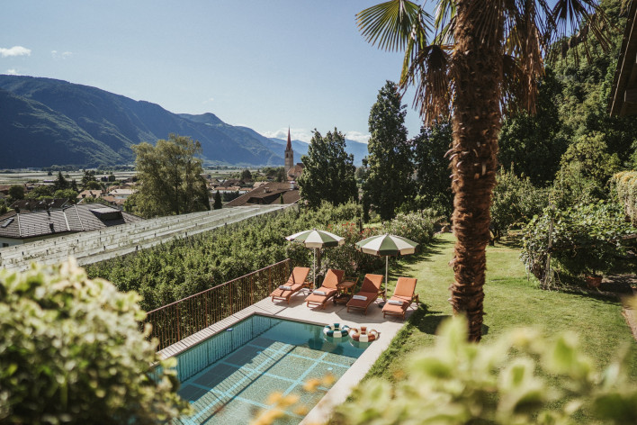 Estate in piscina allo Schnitzbichlhof Piscina con lettini color terracotta, ombrelloni e una palma allo Schnitzbichlhof, con vista su Lana e sulle montagne
