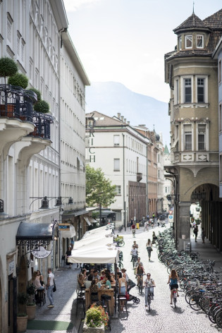 A spasso nel centro storico di Bolzano Via animata di Bolzano con tavolini all’aperto, ciclisti e facciate di edifici storici